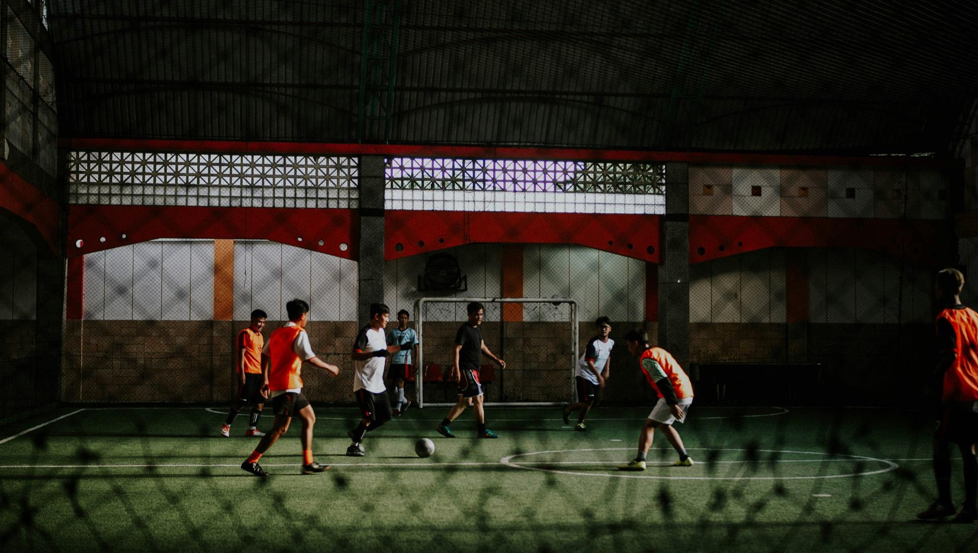 Mejenga de fútbol sala bajo techo, con un equipo en chaleco naranja y el otro de camisa blanca